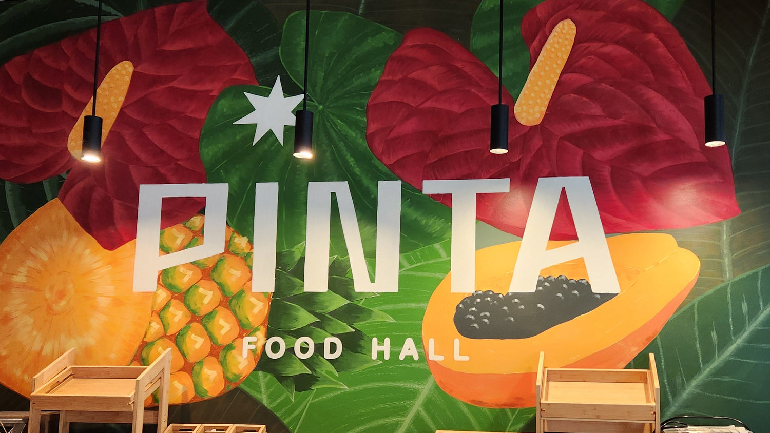 A large wall mural at Pinta Food Hall features tropical fruit and leaf artwork with the words 'PINTA FOOD HALL' in white.